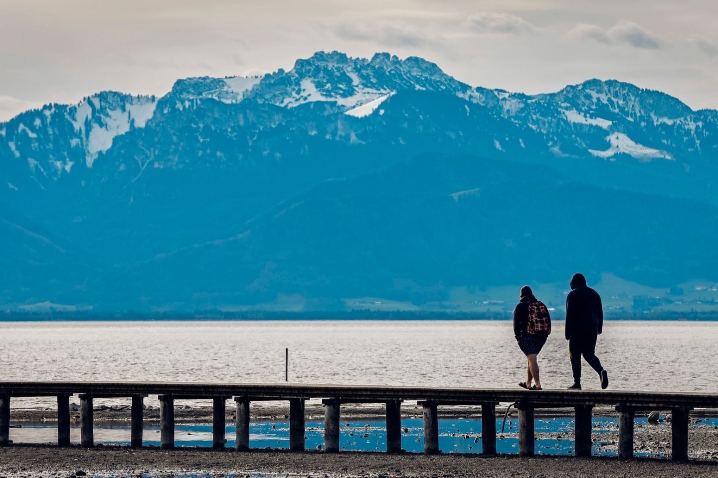 Zwei Männer gehen bei frühlingshaften Temperaturen vor der fast schneefreien Kulisse der Kampenwand in den Chiemgauer Bergen über einen Steg im Chiemsee.